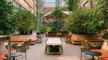 An outdoor patio area with a long table and multiple chairs, surrounded by brick flooring and greenery.