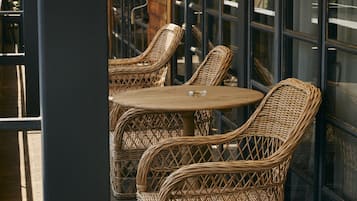 A covered outdoor seating area with wicker chairs and tables, large windows, and a brick wall.