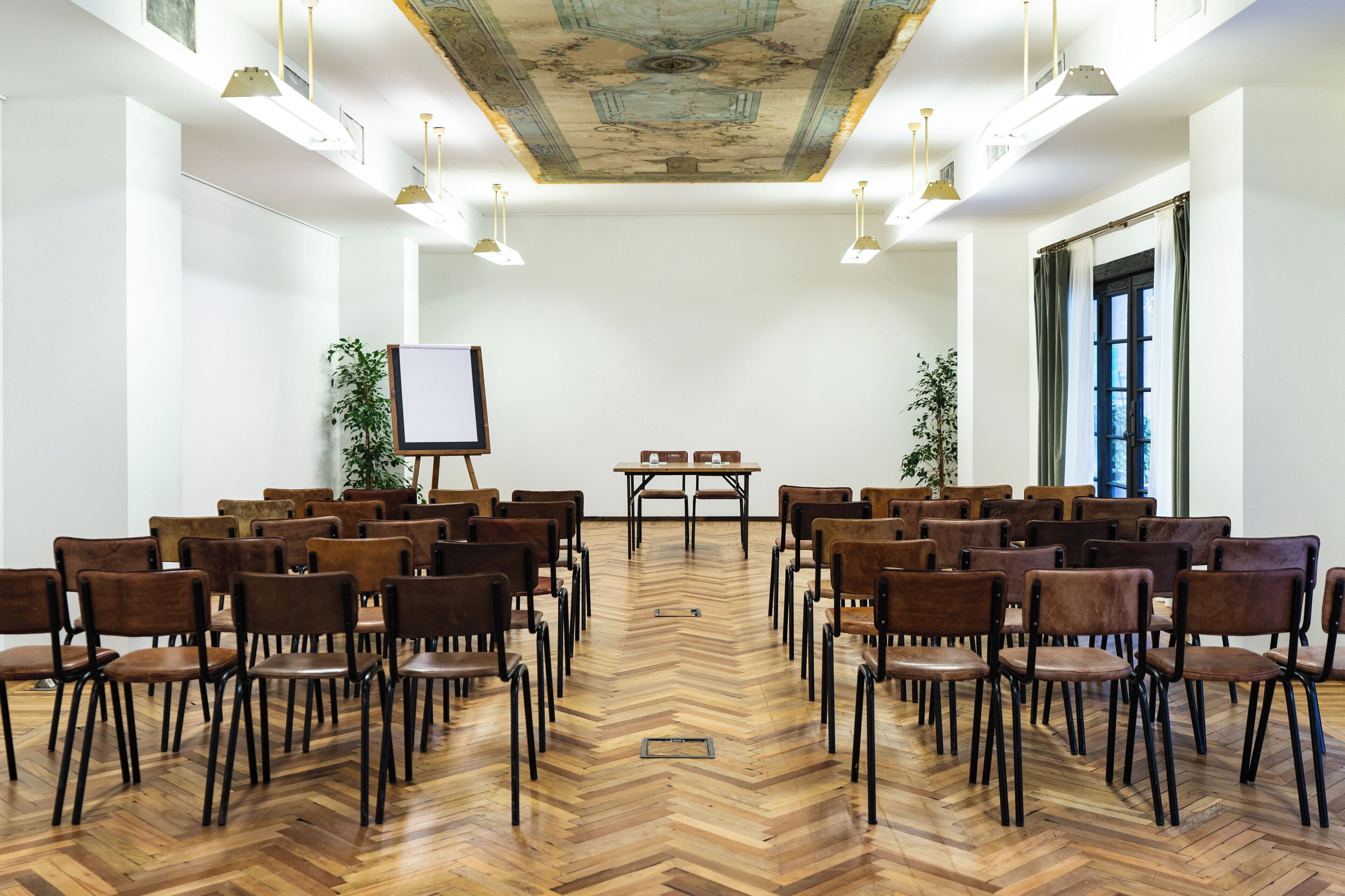 A conference room with a wooden herringbone floor, rows of brown chairs, a whiteboard, and a table with chairs.
