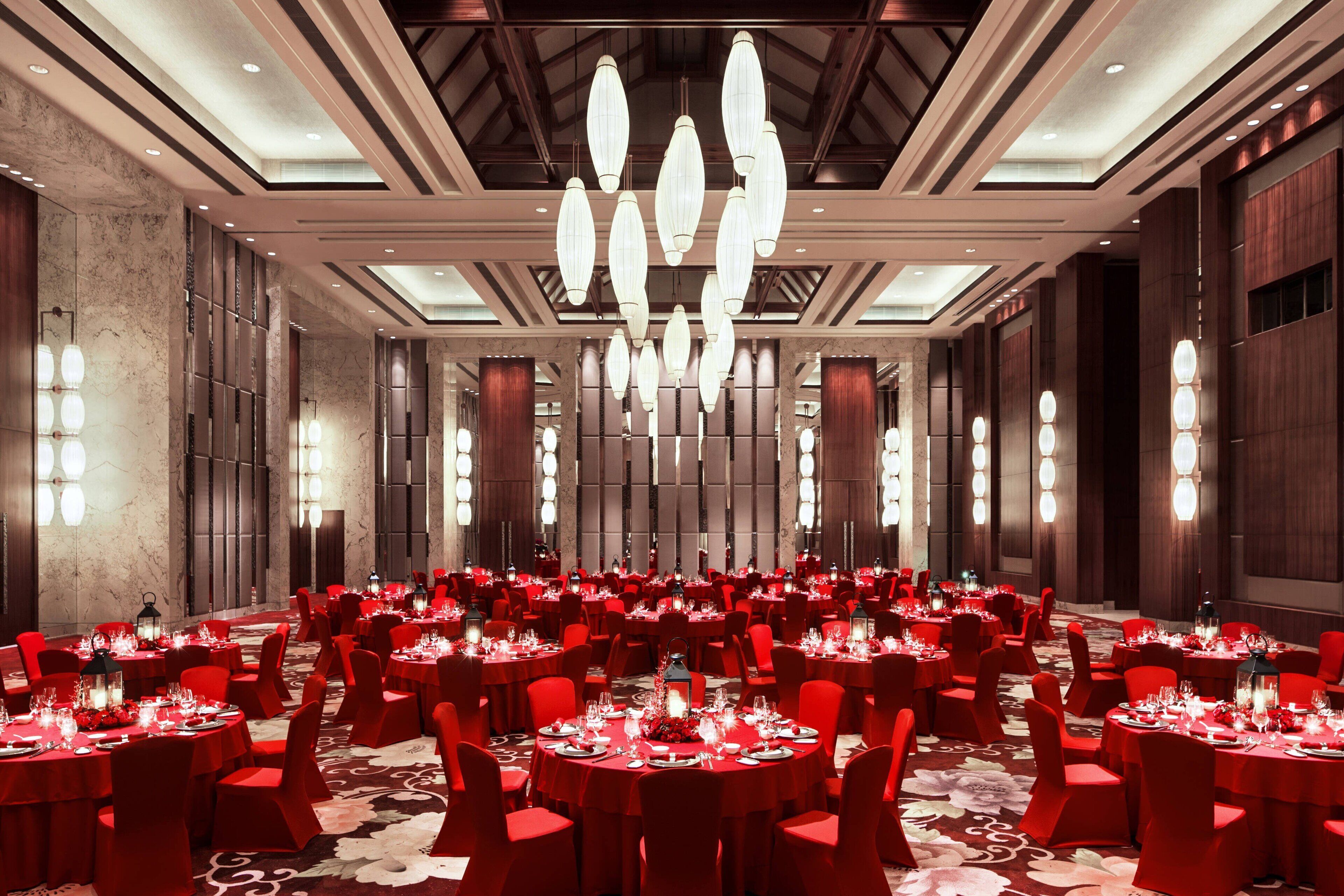 A large banquet hall with round tables set for dining, red chairs, and chandeliers.
