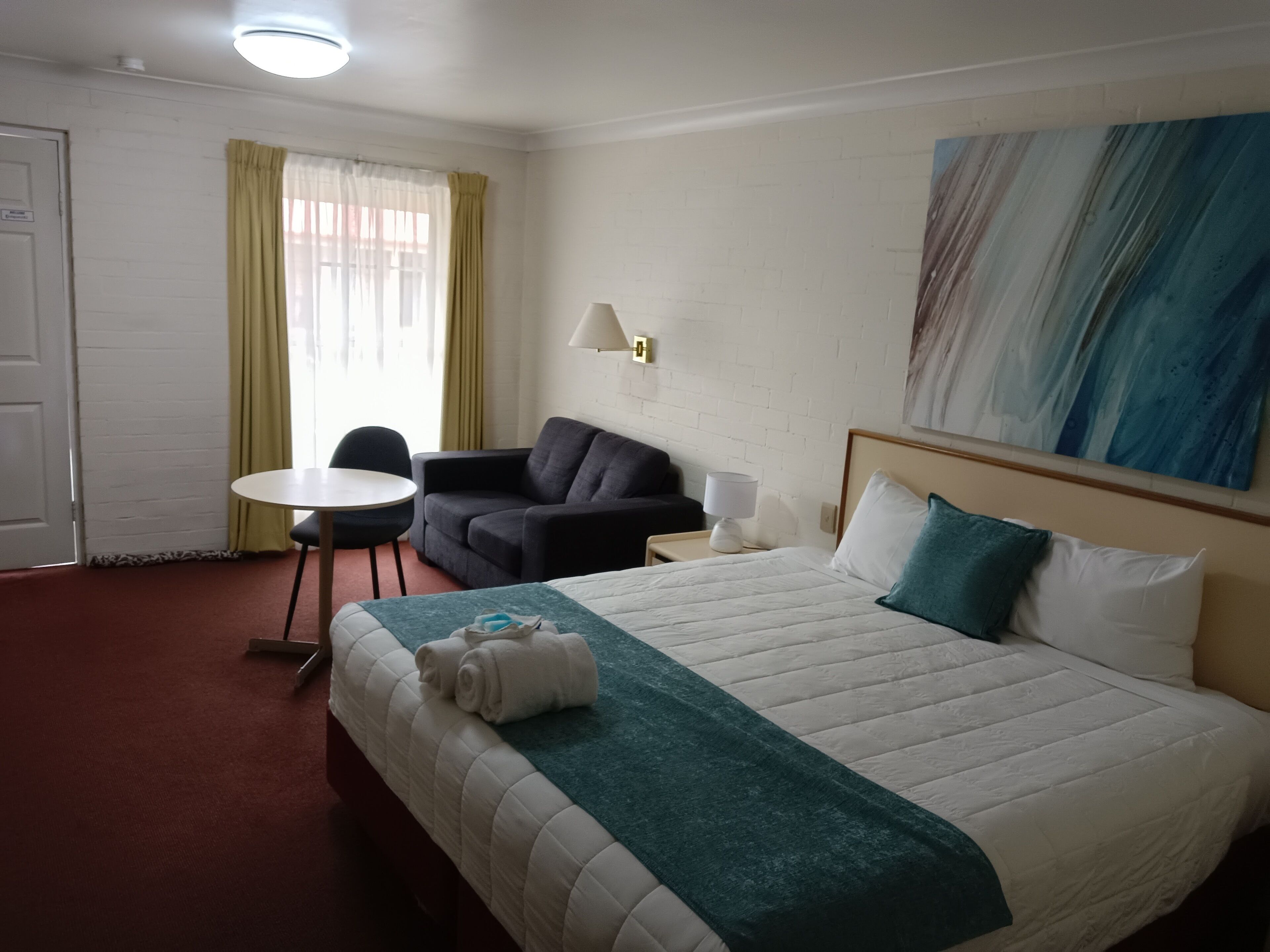 A hotel room with a bed, a desk, a chair, a sofa, a lamp, and a painting on the wall.