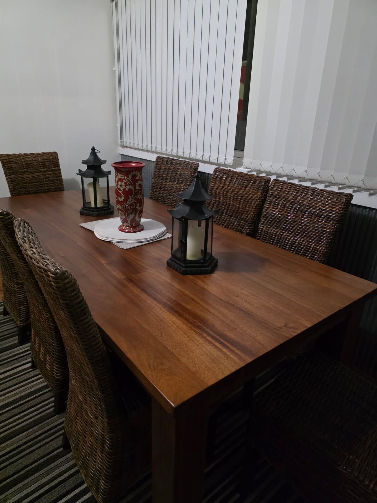 A dining table with wicker chairs, two black lanterns, and a red vase.