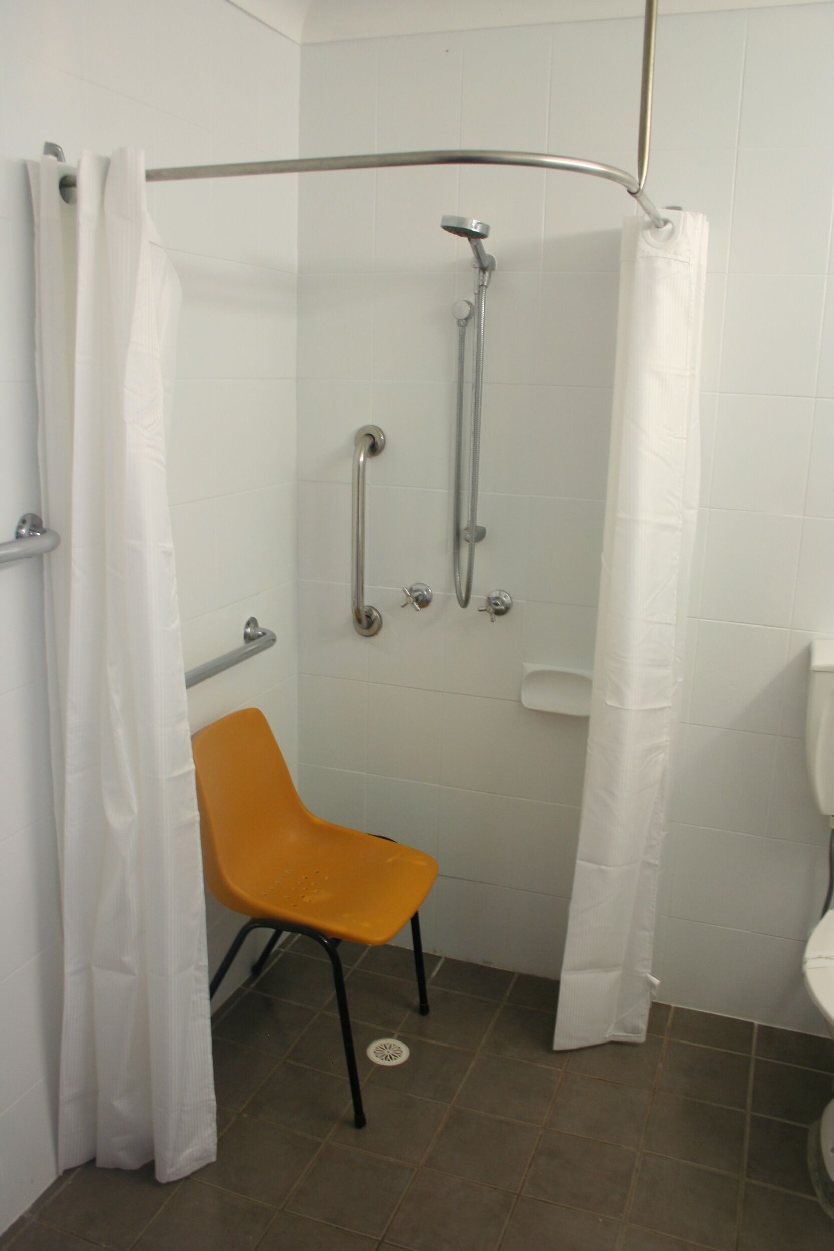 A shower area with a white shower curtain, a grab bar, a handheld showerhead, and a yellow chair.