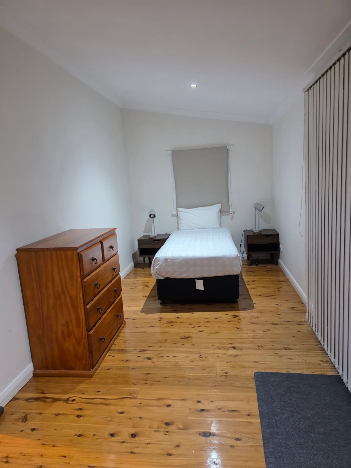 A bedroom with a wooden dresser, a bed with a white mattress, two bedside tables with lamps, and a window with a blind.