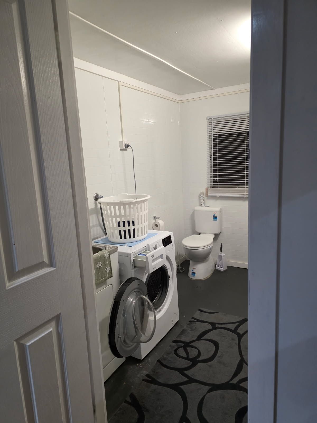 A laundry room with a front-loading washing machine, a white toilet, and a window with blinds.
