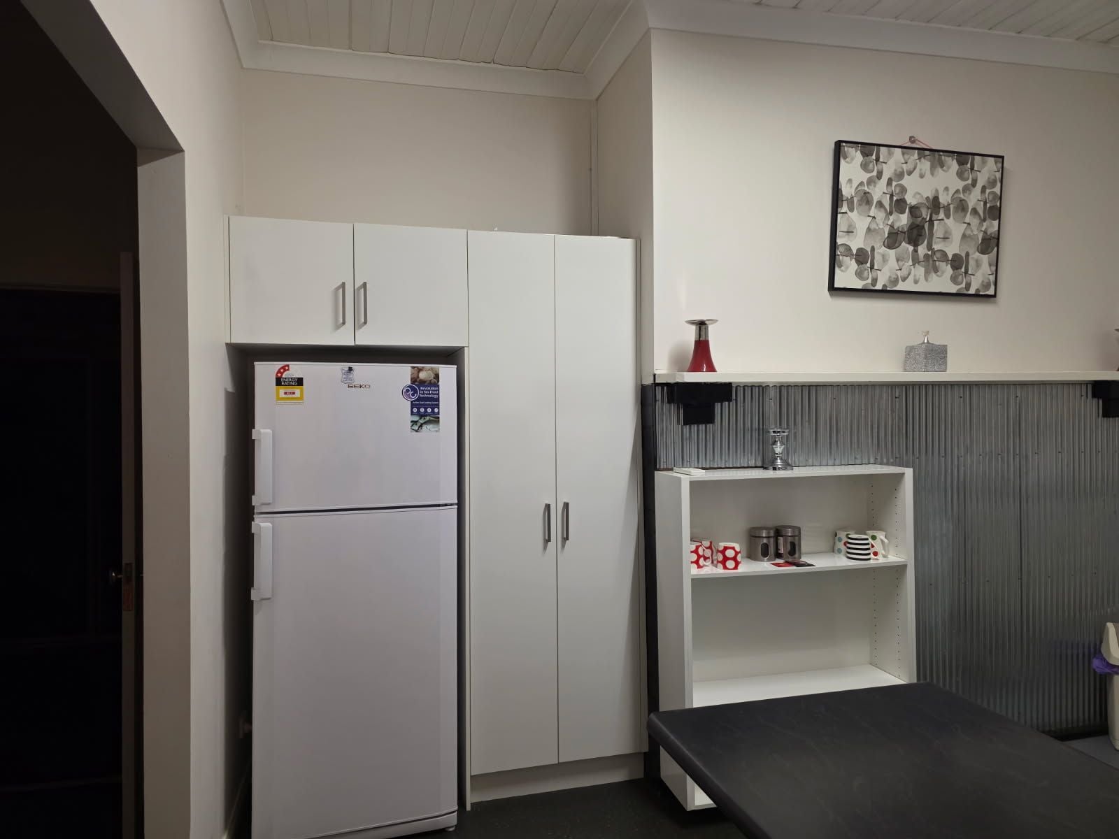 Una kitchenette con un refrigerador blanco, una repisa con utensilios de cocina y un cuadro colgado en la pared.