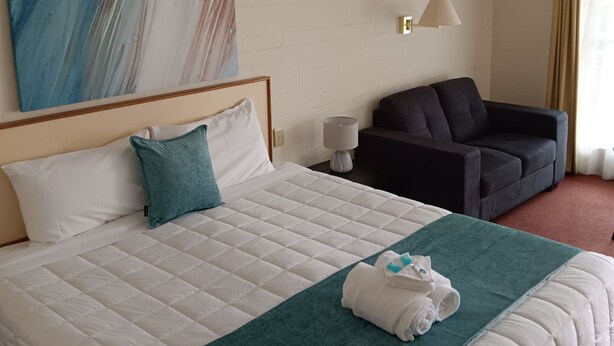 Habitación de hotel con una cama grande, un sillón oscuro, una mesita con lámpara y un cuadro en la pared.