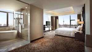 A hotel room with a large bed, a separate bathroom with a bathtub, a desk with a TV, and a view of a city skyline. - Ramada Plaza Xian South (Xi'an)