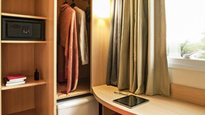 A compact room with a wooden desk, a red chair, a small refrigerator, a shelf with books and a bottle, and a window with curtains. - Ibis Rio de Janeiro Parque Olimpico (Rio de Janeiro)
