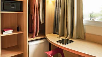 Une pièce de taille modeste, avec un bureau en bois, une chaise rouge, un petit réfrigérateur, une étagère avec des livres et une bouteille, et une fenêtre avec des rideaux.