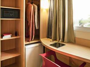 A compact room with a wooden desk, a red chair, a small refrigerator, a shelf with books and a bottle, and a window with curtains. - Ibis Rio de Janeiro Parque Olimpico (Rio de Janeiro)