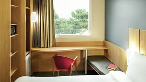 A hotel room with a bed, a desk with a chair, a window with curtains, and a bookshelf. - Ibis Rio de Janeiro Parque Olimpico (Rio de Janeiro)