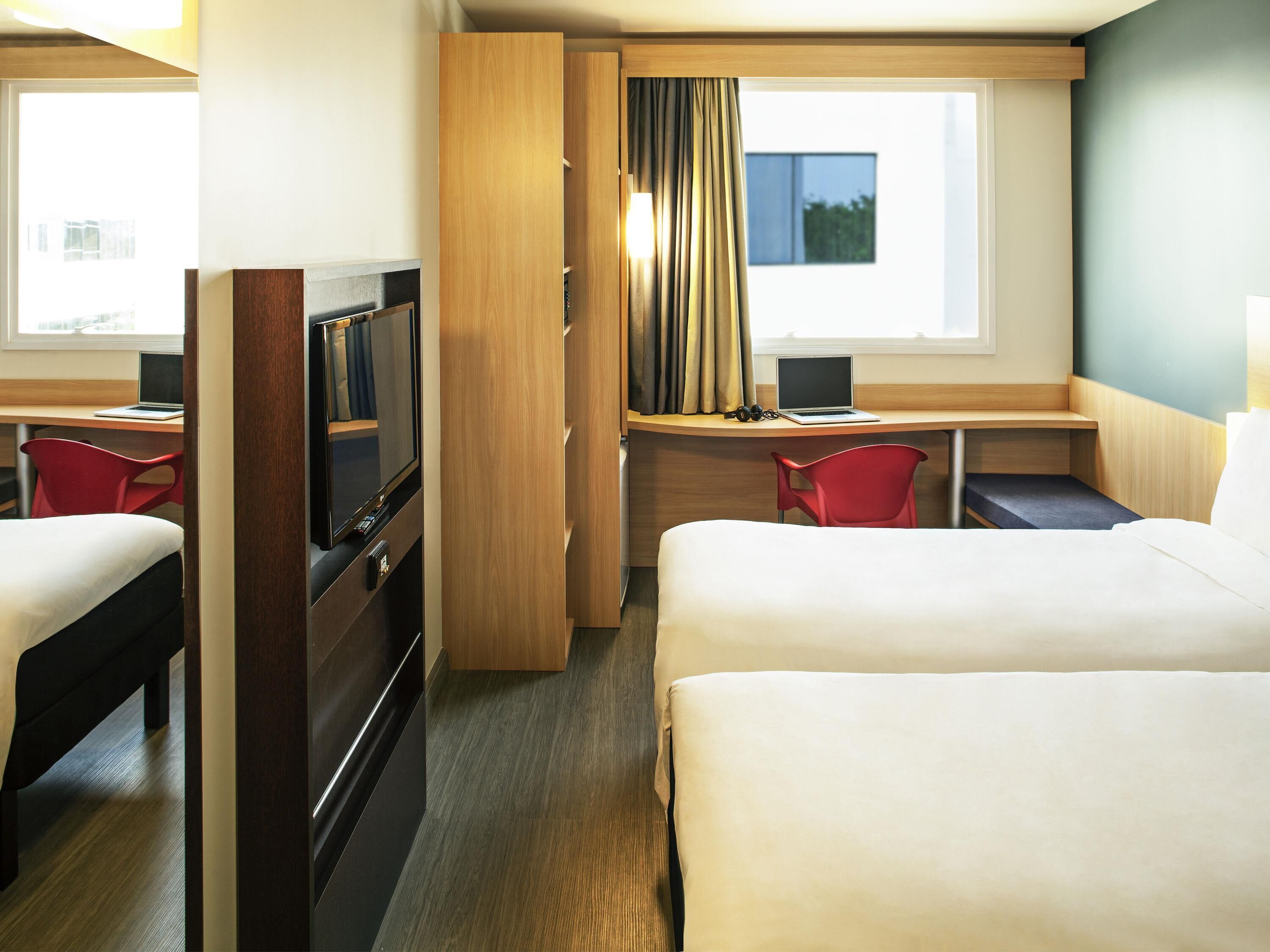A hotel room with two beds, a desk with a laptop, a TV, and a red chair.