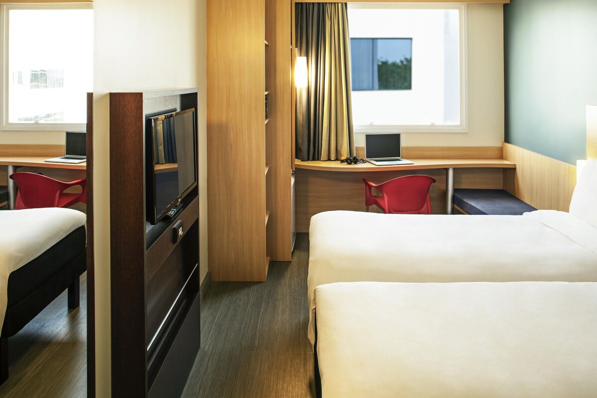A hotel room with two beds, a desk with a laptop, a TV, and a red chair.