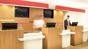 A reception area with wooden reception desks, a flat-screen TV mounted on the wall, and a person standing in the background. - Ibis Rio de Janeiro Parque Olimpico (Rio de Janeiro)