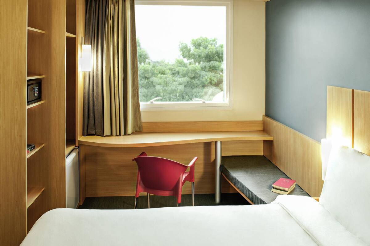 A hotel room with a bed, a desk with a chair, a window with curtains, and a bookshelf.