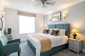 A neatly arranged bedroom with a bed, bedside tables, a chair, a desk, and a ceiling fan. - SACO Jersey Merlin House (St. Helier)