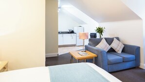 Una habitación compacta con una cama, un sofá, una mesita y una lámpara, con una zona de cocina al fondo.