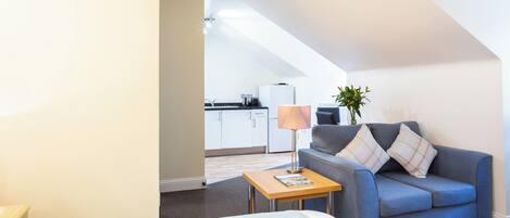 A compact room with a bed, a sofa, a small table, a lamp, and a kitchen area in the background.