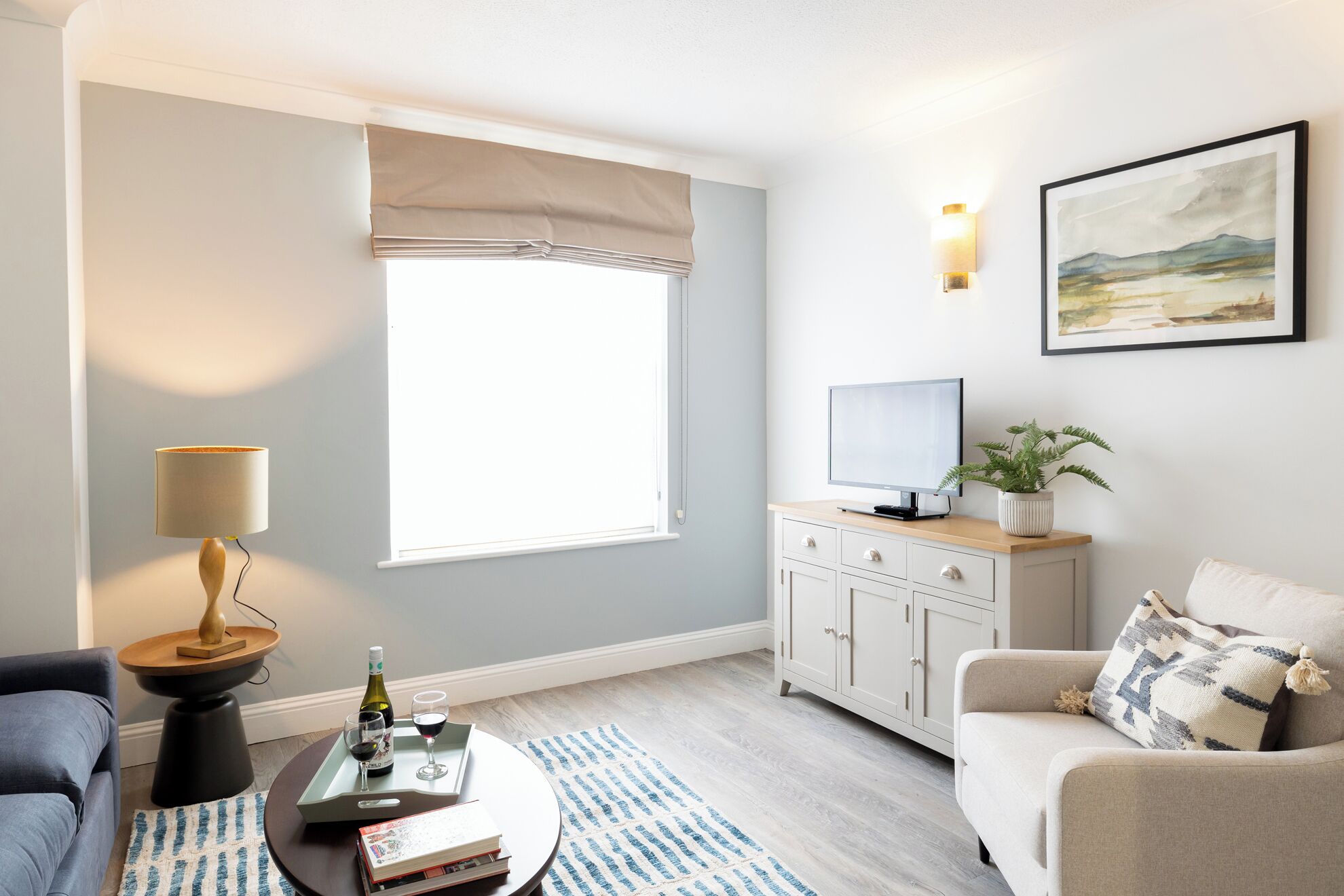 Una sala de estar con un sofá, una mesa de centro con libros y una botella, un televisor en un mueble, una ventana con persiana y un cuadro en la pared.