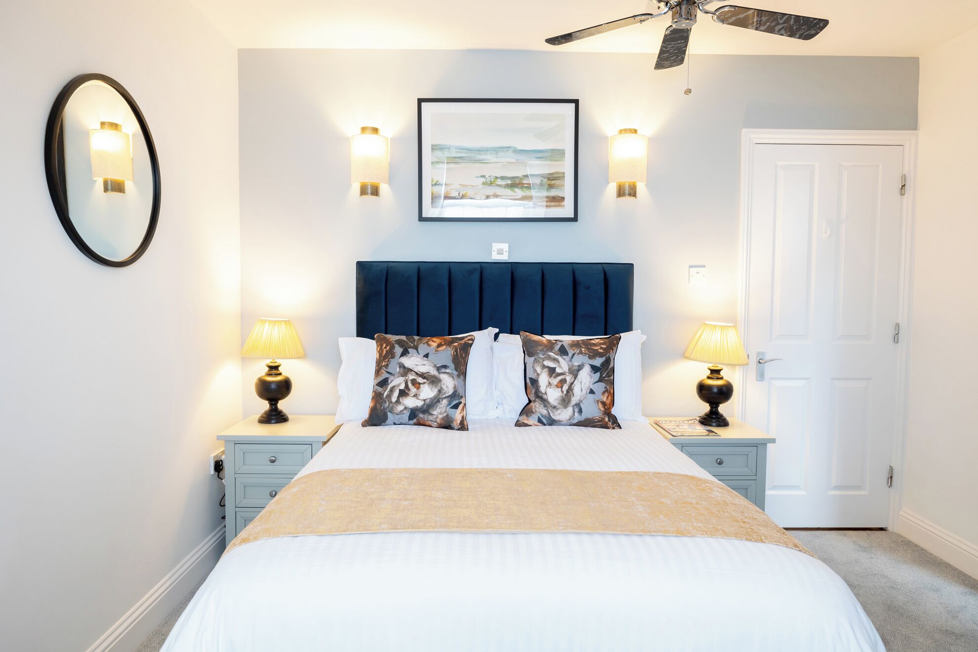 A bedroom with a bed, two bedside tables, a ceiling fan, a framed picture, and a door.