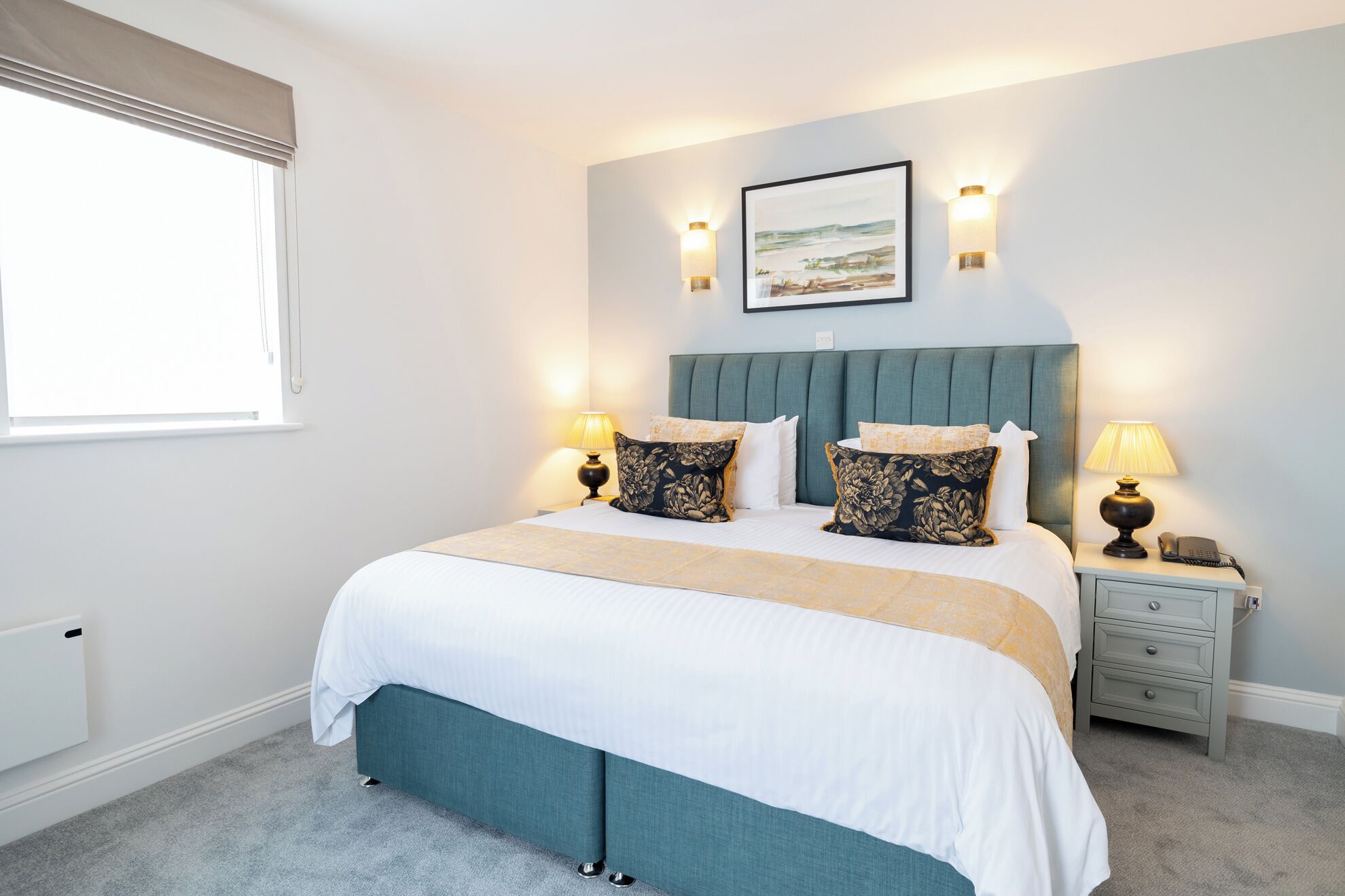 A bedroom with a bed, two bedside lamps, a nightstand, a framed picture on the wall, and a window with blinds.