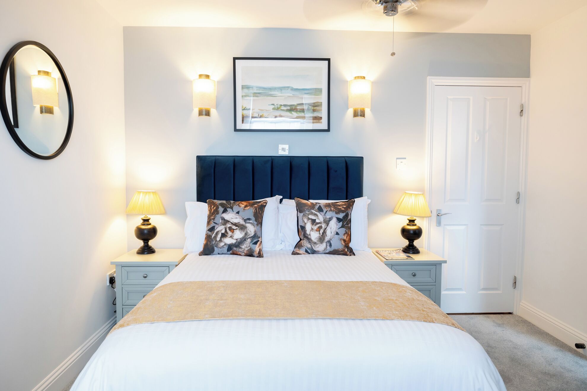 A bedroom with a bed, two bedside lamps, a framed picture on the wall, and a door.