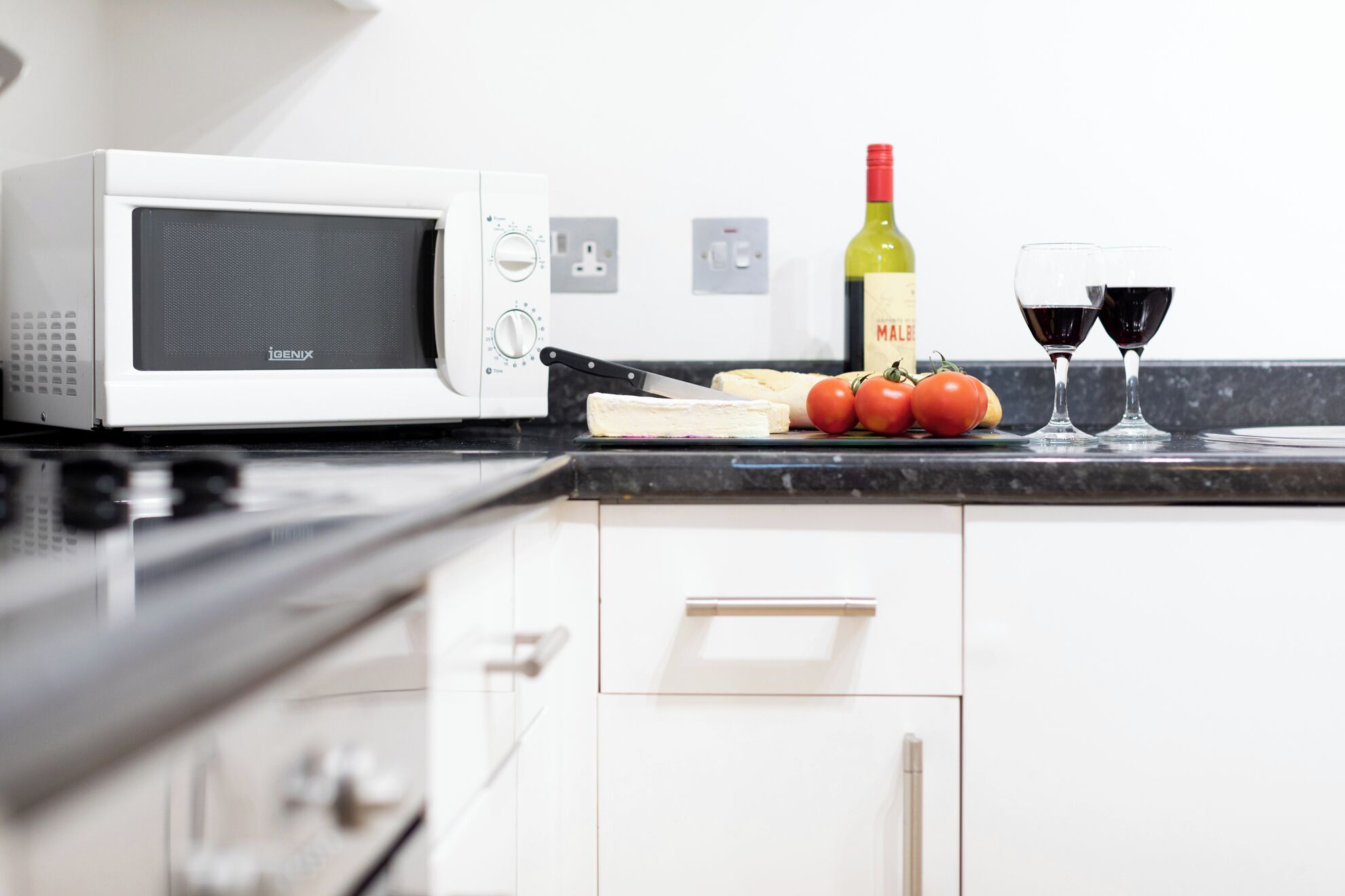 A kitchen with a white microwave, a bottle of wine, and two glasses on the counter.
