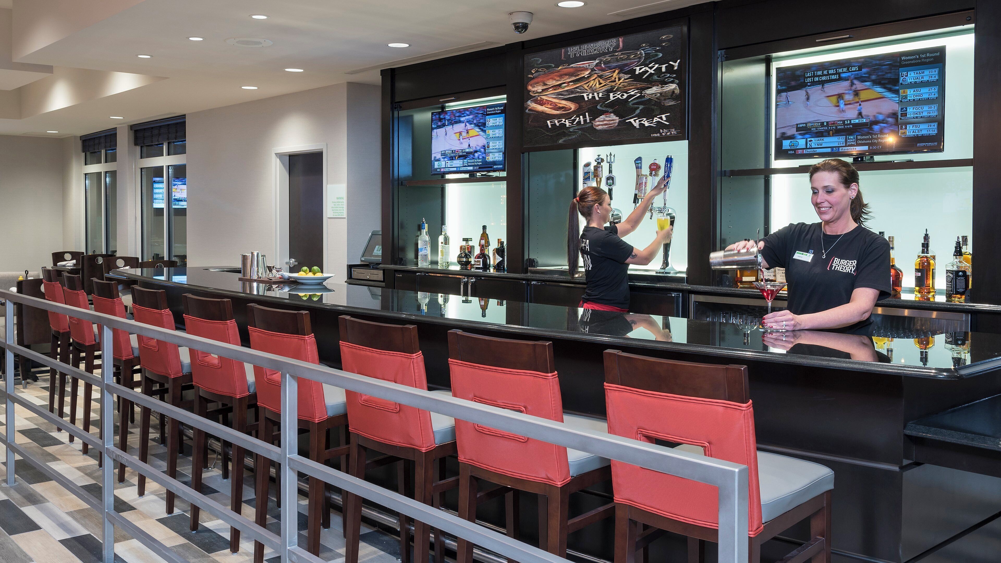 A bar area with a long counter, multiple seating options, and a television screen displaying sports.