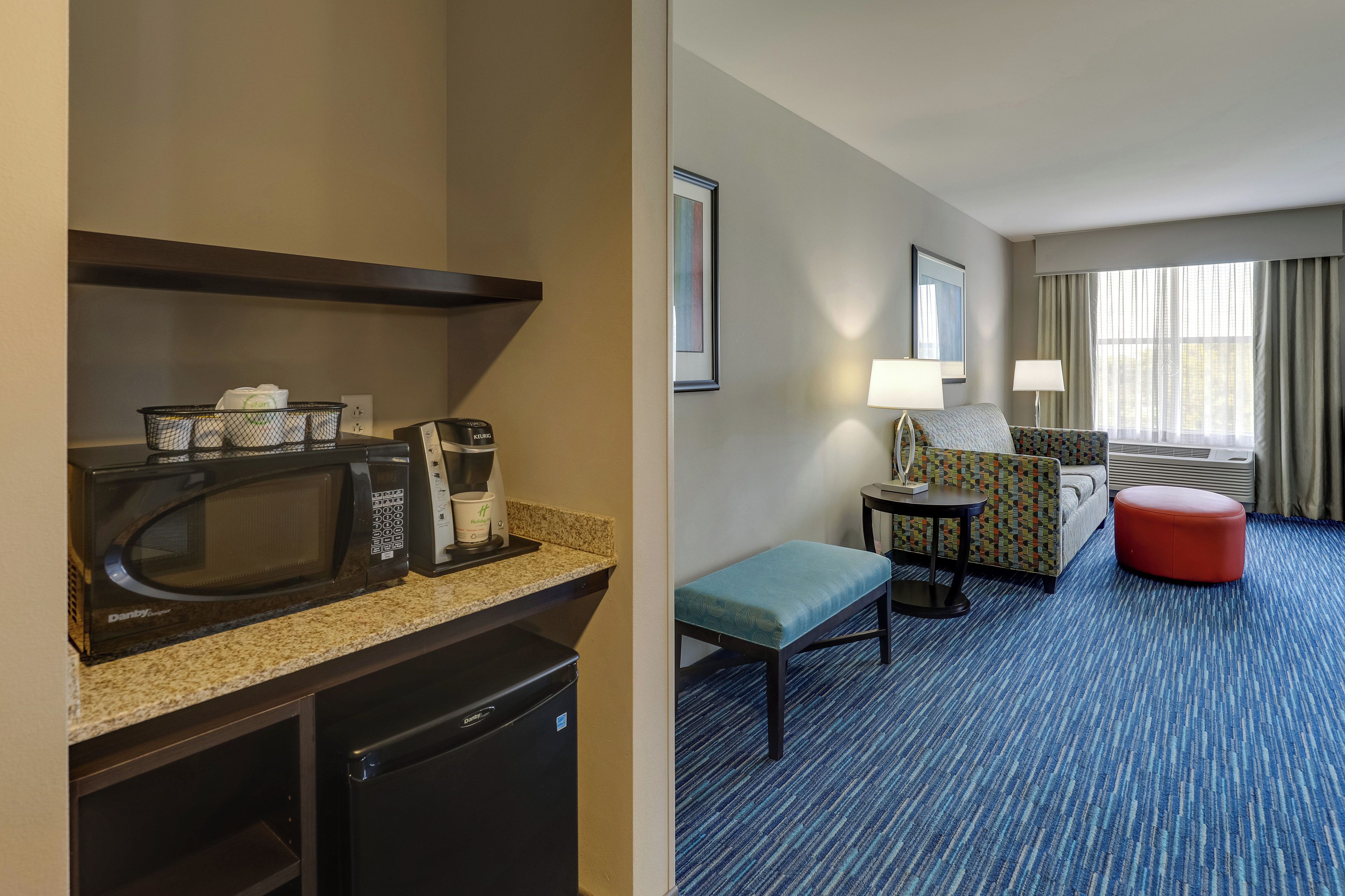 A hotel room with a kitchenette, a seating area with a sofa and ottoman, and a window with curtains.