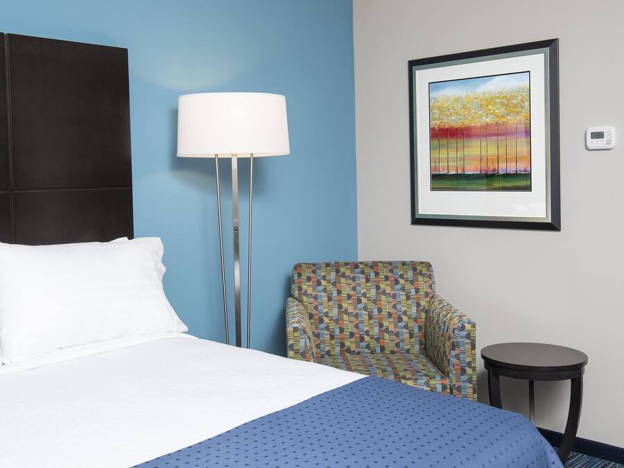 Una habitación de hotel con una cama, una silla, una lámpara, un cuadro enmarcado y una puerta.