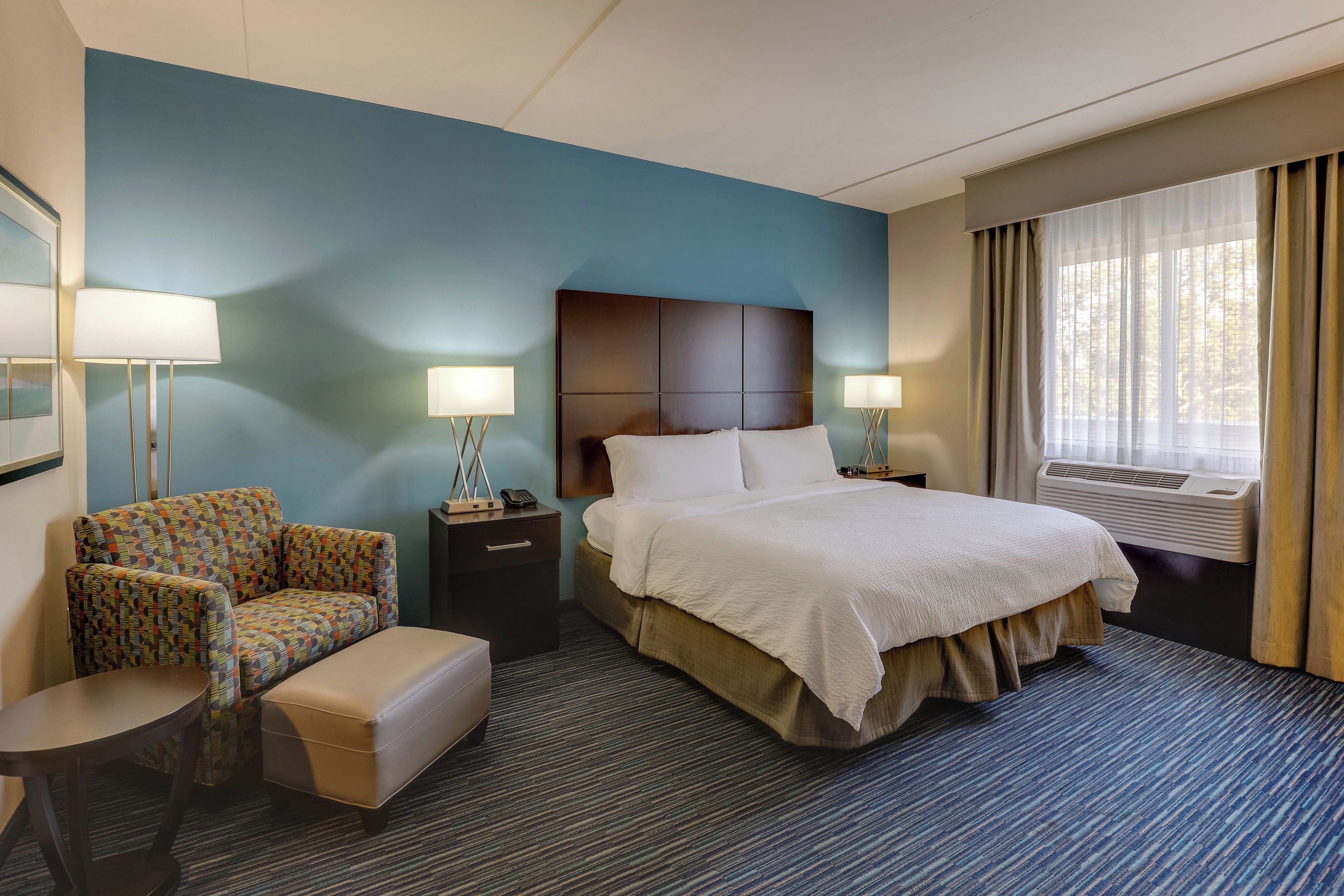 A hotel room with a large bed, two bedside tables, a chair, a small table, and a window with curtains.