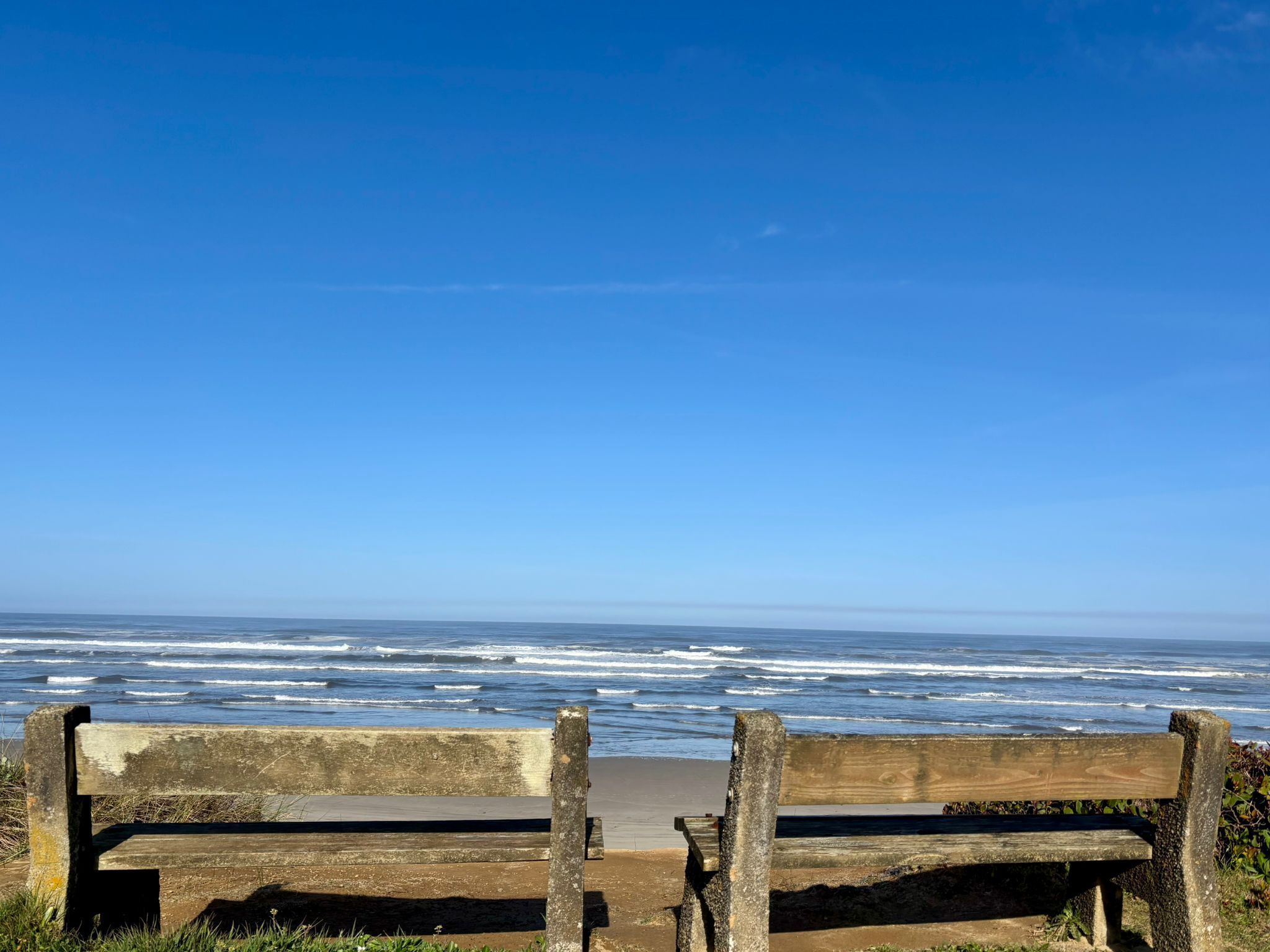 Two wooden benches facing the ocean with waves crashing onto the shore under a clear blue sky.