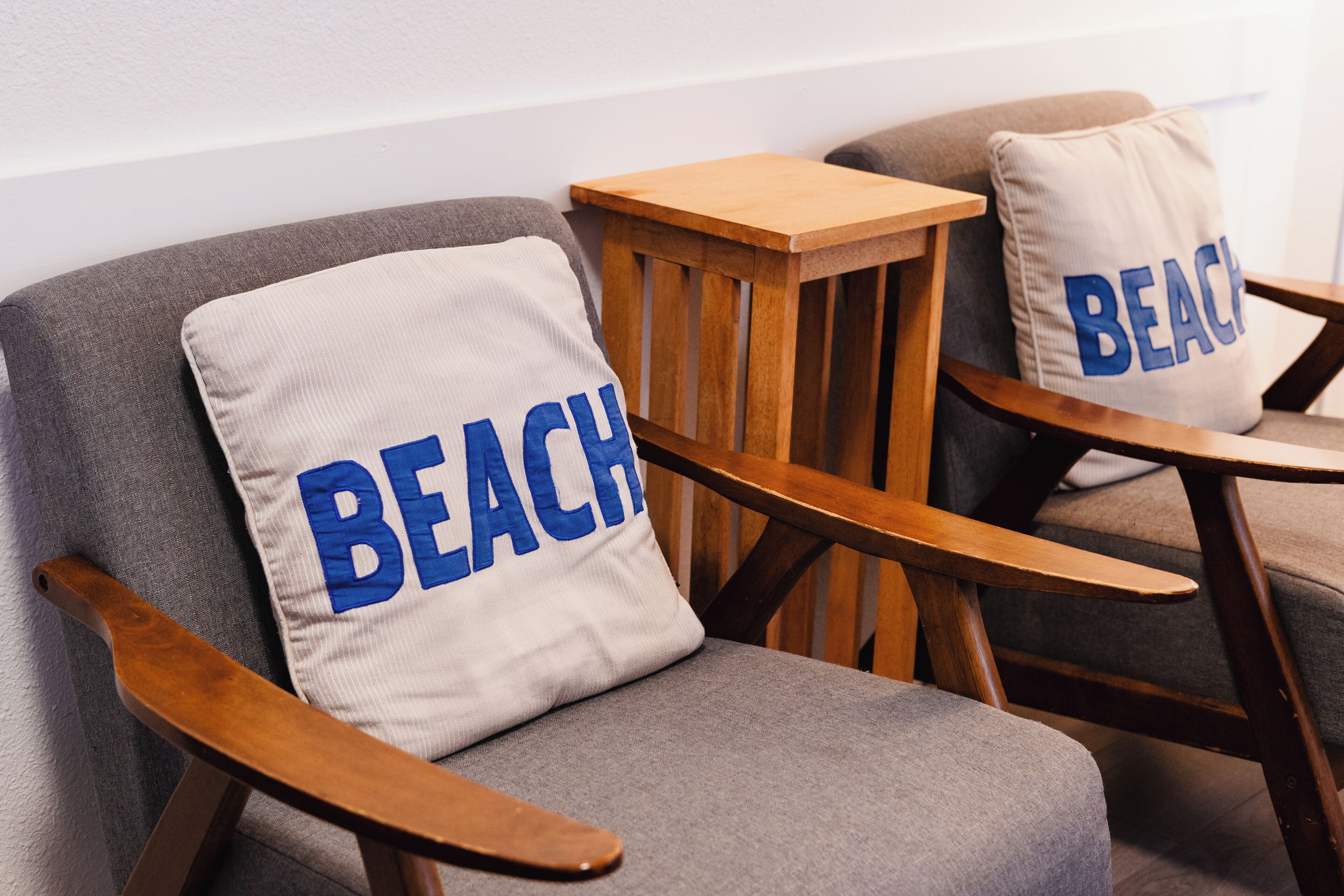 Two wooden chairs with grey upholstery and pillows with the word 'BEACH' on them.