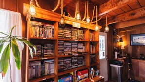 Una habitación con estanterías de madera llenas de libros y DVD, un cesto de basura pequeño, una planta en maceta y un televisor en la pared.