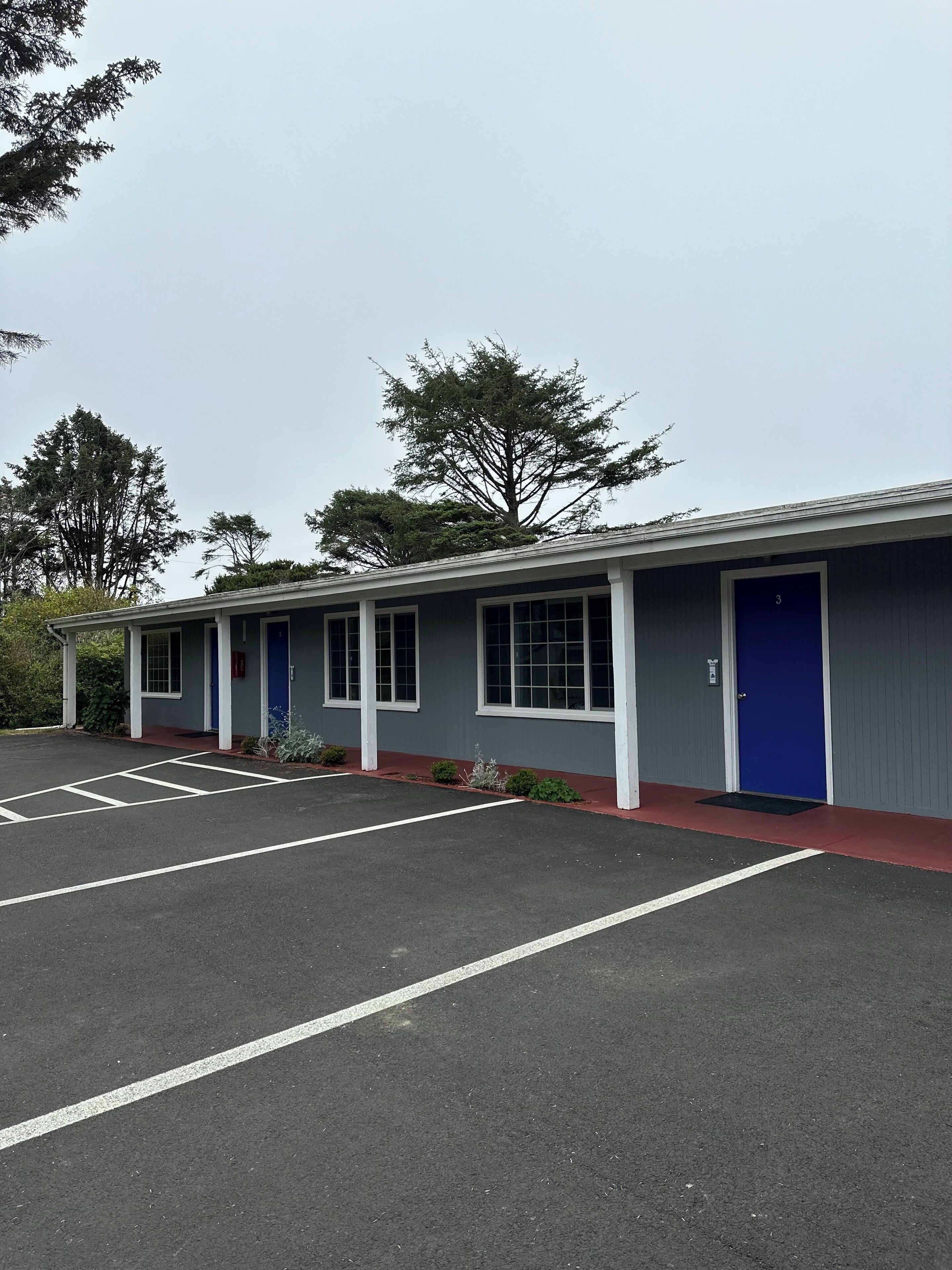 A motel with a row of rooms, each with a blue door and a window. There is a parking lot in front with marked spaces.