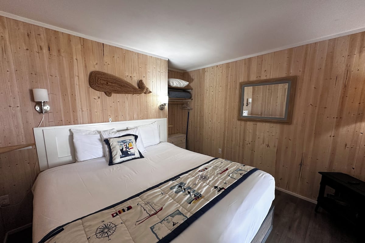 A bedroom with a wooden paneled wall, a bed with a nautical-themed duvet, a wall-mounted lamp, and a mirror.