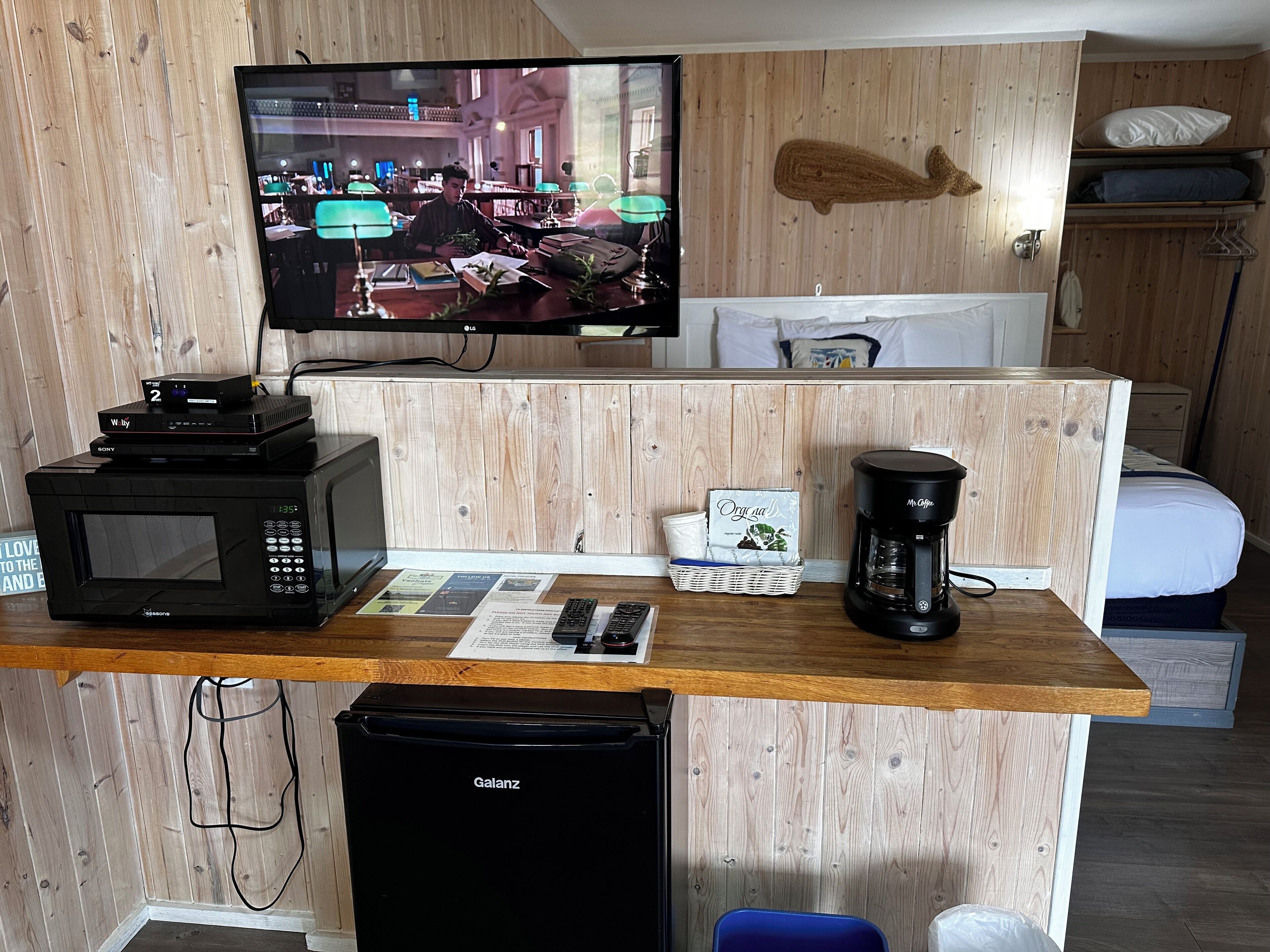 Habitación de hotel compacta con detalles de madera, un televisor de pantalla plana en la pared, microondas, cafetera y un refrigerador pequeño.
