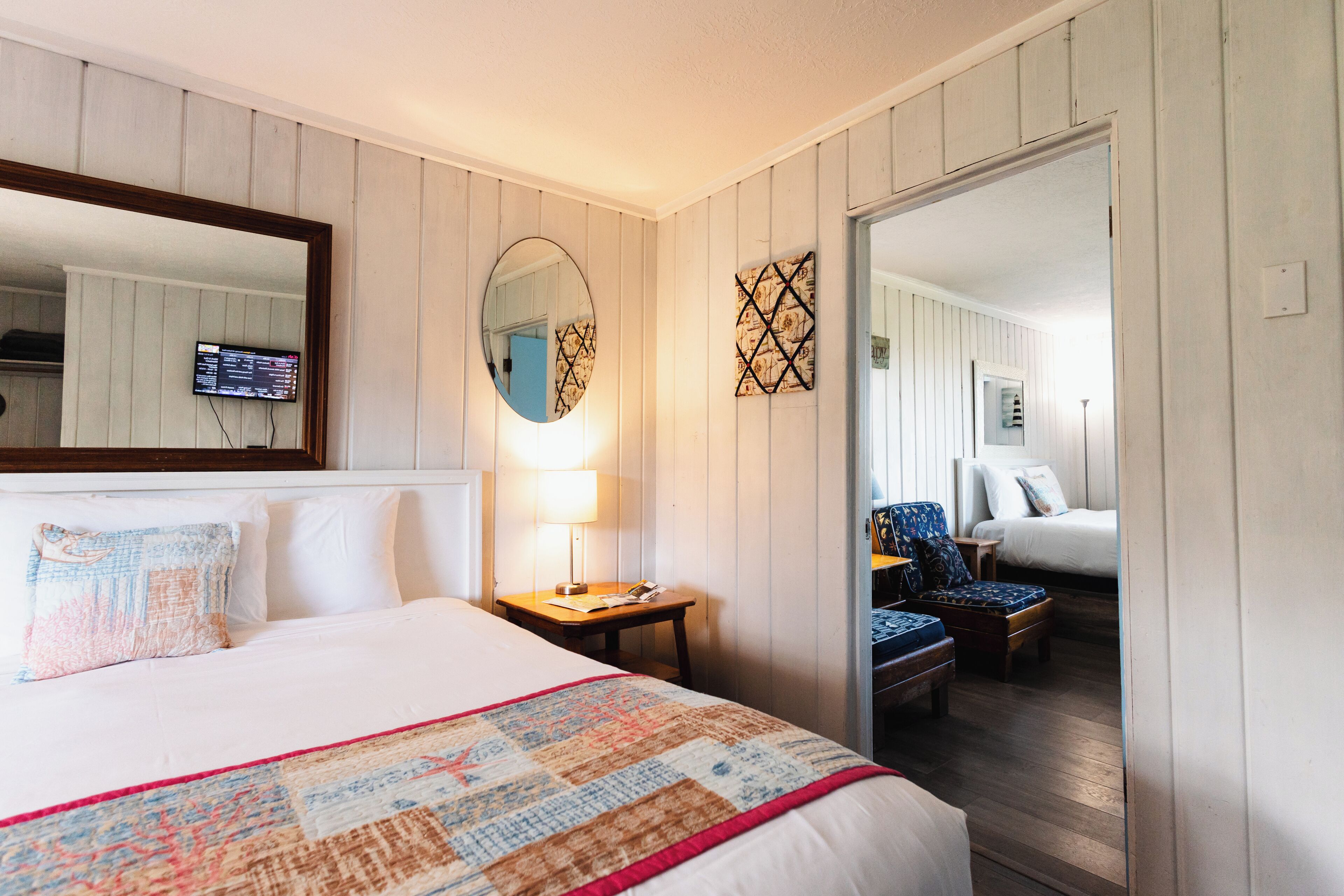 Een hotelkamer met een bed, een nachtkastje met een lamp, een spiegel, een aan de muur bevestigde televisie, en een deur die naar een andere kamer leidt.