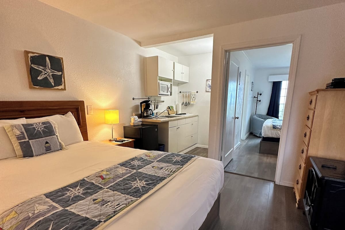 A hotel room with a bed, a desk, a kitchenette, and a bathroom visible through an open door.