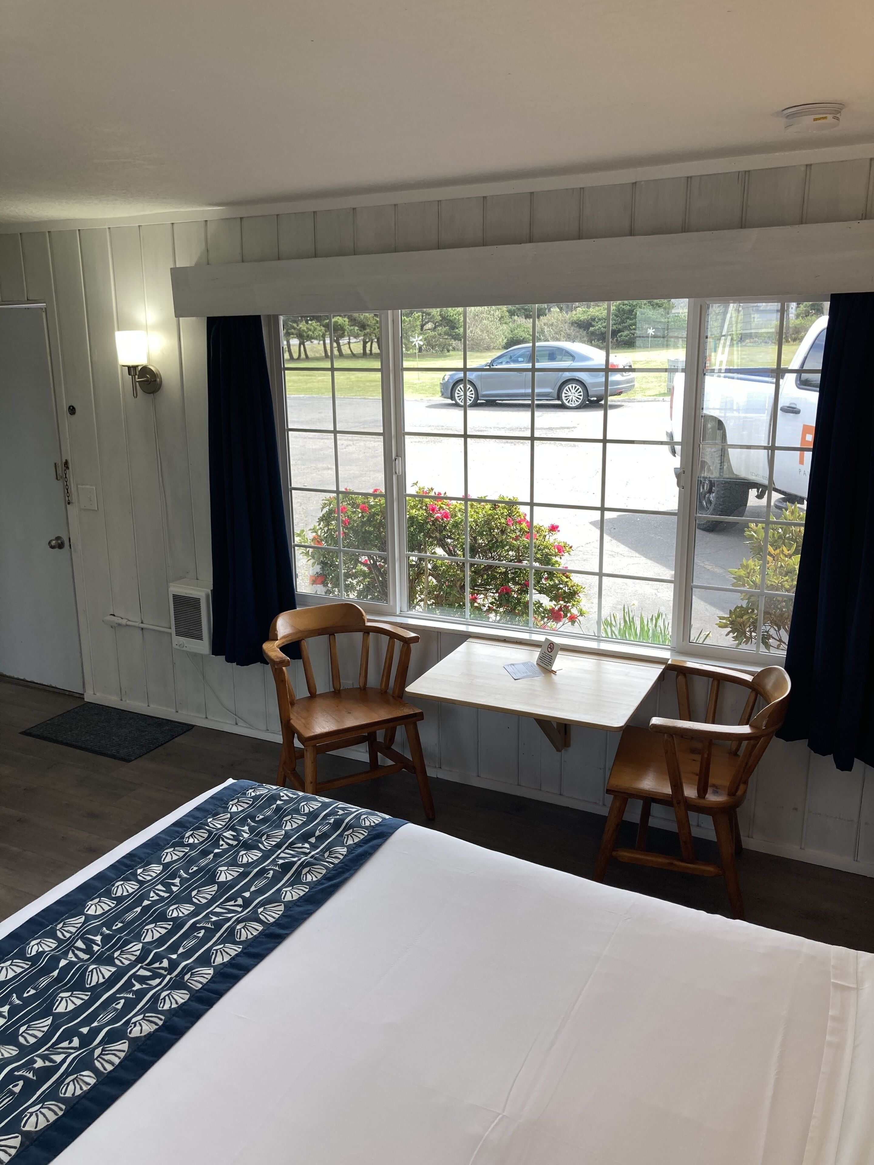 A room with a bed, two wooden chairs, a small table, and a window with a view of parked cars and greenery outside.