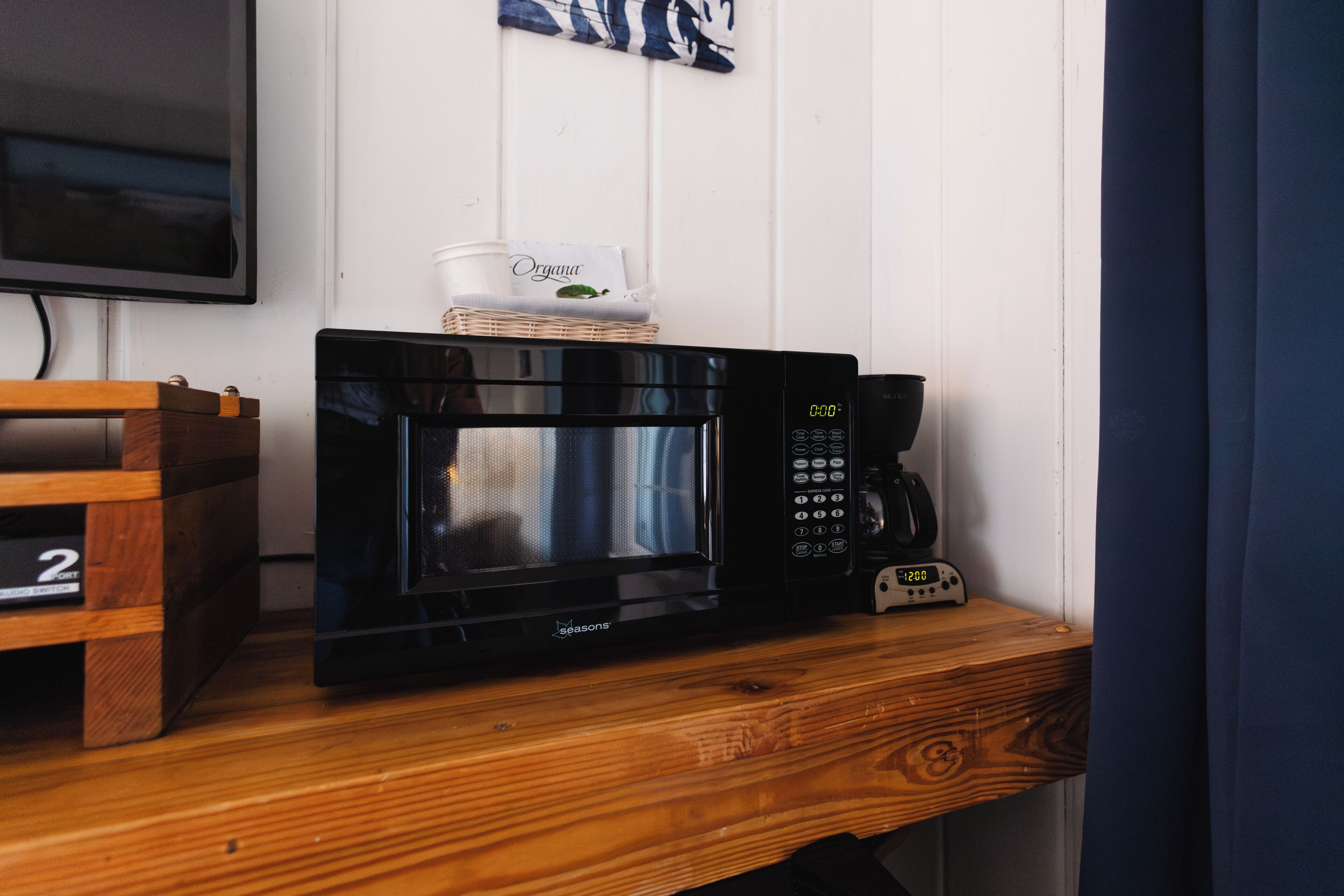 A microwave oven on a wooden shelf, a basket with a coffee cup, and a digital clock displaying 12:08.