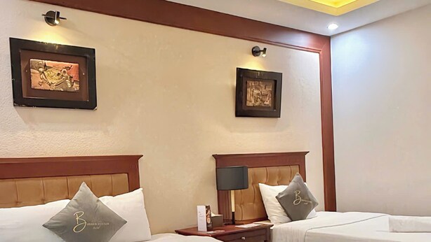Habitación de hotel con dos camas, cabeceros de madera, una lámpara en el techo, obras de arte enmarcadas en las paredes y piso de madera.