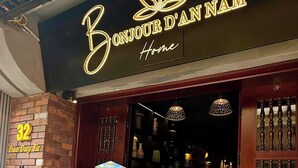Una fachada con un letrero de neón que dice 'Bonjour d'Annam Home'. Hay dos paneles informativos afuera, un muro de ladrillo con los números '32' y 'Dào Dụy Tự', y una planta en maceta a la izquierda.