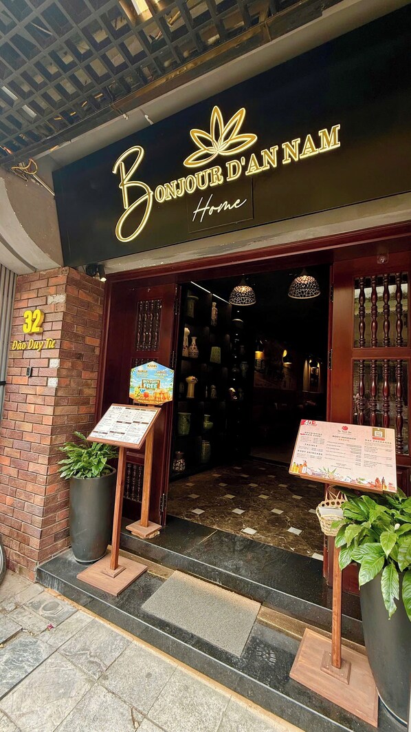 A storefront with a neon sign reading 'Bonjour d'Annam Home.' There are two informational boards outside, a brick wall with the number '32' and 'Dào Dụy Tự,' and a potted plant on the left. - Bonjour D'An Nam Home (Hanoi)