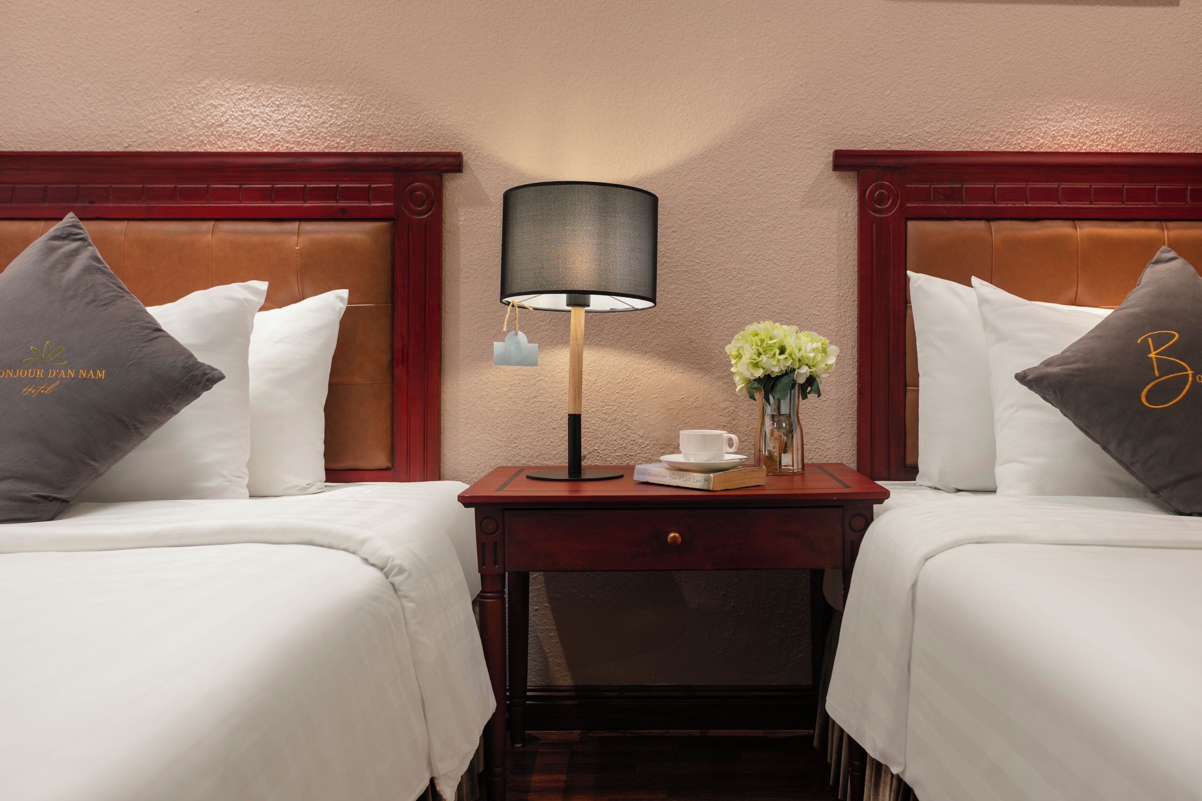 A hotel room with two beds, a nightstand with a lamp, a vase with flowers, and a cup.