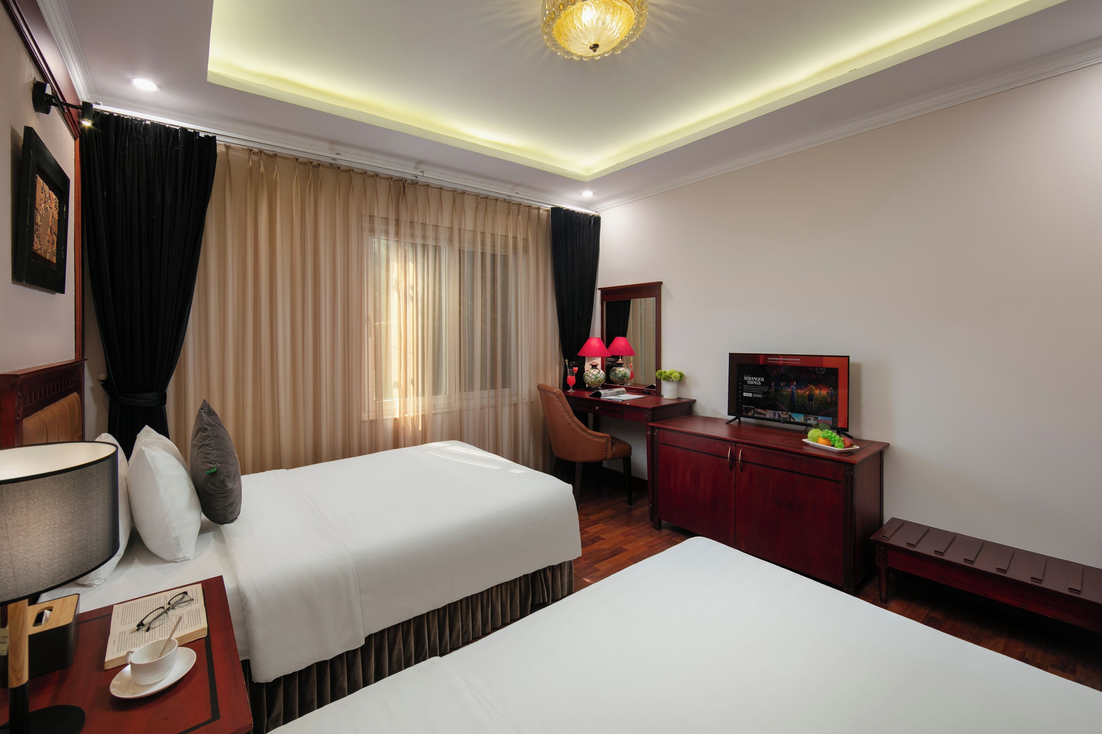 A hotel room with two beds, a desk, a TV, and a bench.