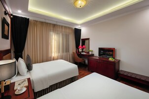A hotel room with two beds, a desk, a TV, and a bench.