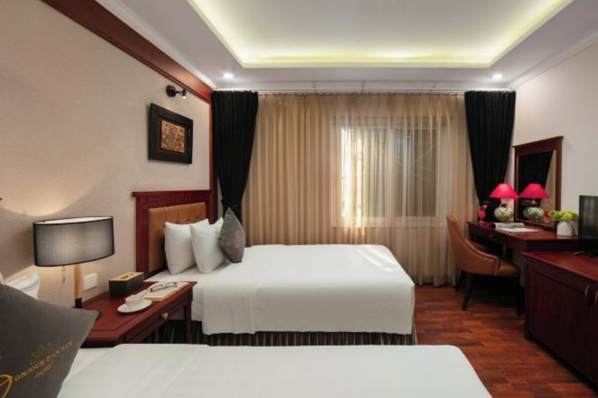 Quarto de hotel com cama, mesa de cabeceira, luminária, escrivaninha, cadeira e janela com cortinas.