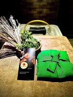 Una cesta de mimbre llena de hierbas verdes, un paño verde atado con un lazo y una tarjeta negra con el logotipo y texto de un spa.